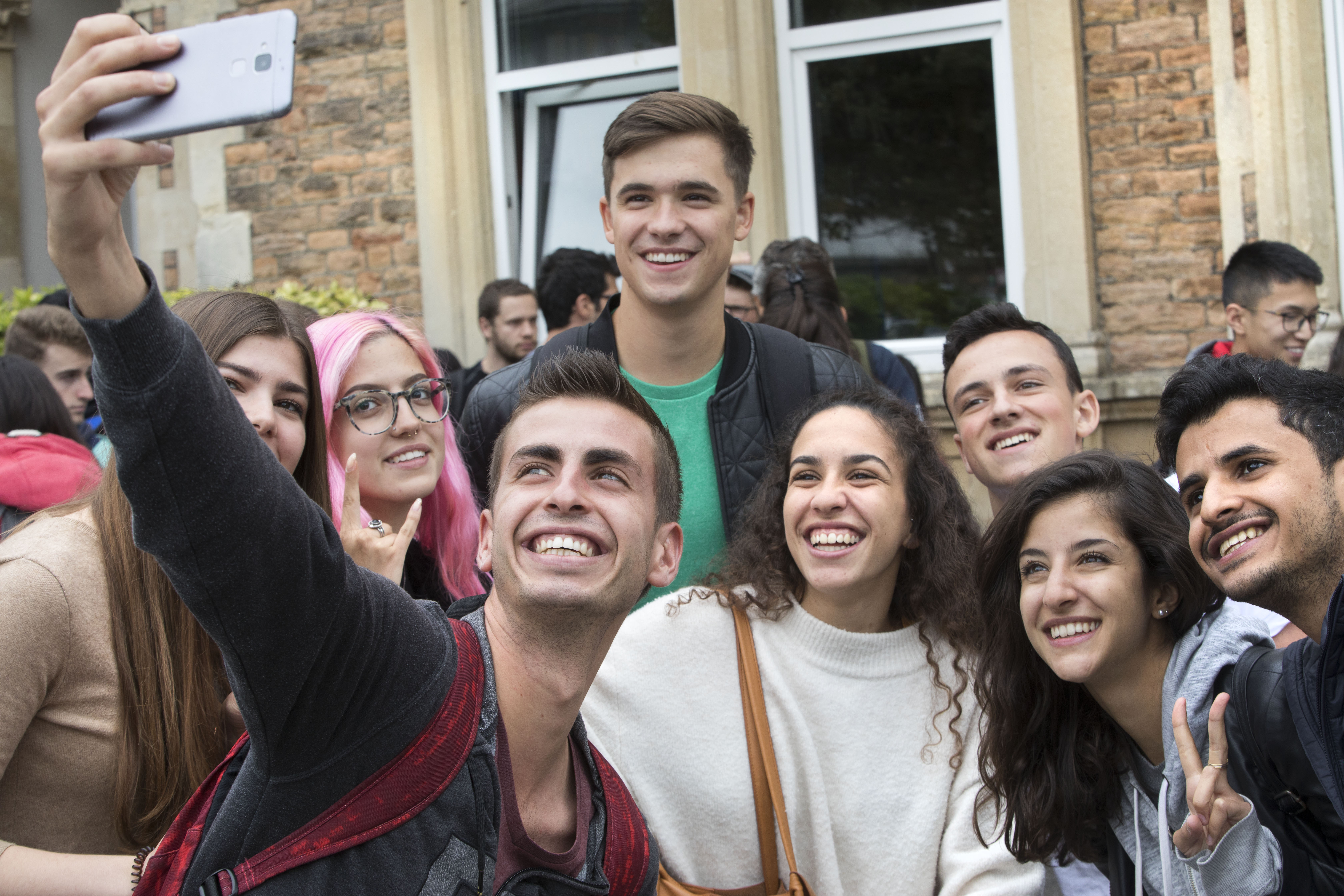Group of Young adults taking a selfie, crouched down in front of the school building.
