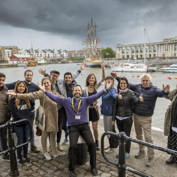 Group of students stood with their hands in the air, with Bristol dockside in the background